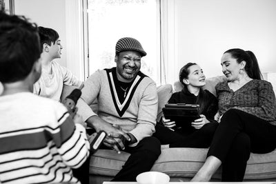 A family sits together on a couch, smiling and laughing. Two children face each other, one holding a tablet. An adult man in a hat sits in the center, laughing, while a woman and a girl talk and smile on the right.