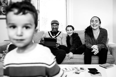 A black and white photo of a family in a living room. A young boy is in the foreground, out of focus. In the background, three adults sit on a couch smiling, with a window behind them and cards on the table.