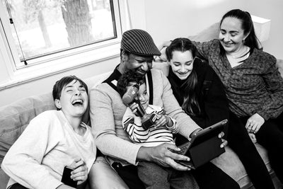 A family of five sits closely on a couch, smiling and laughing together while looking at a tablet. The youngest child holds a toy and sits on an adults lap. The mood is joyful and lively.