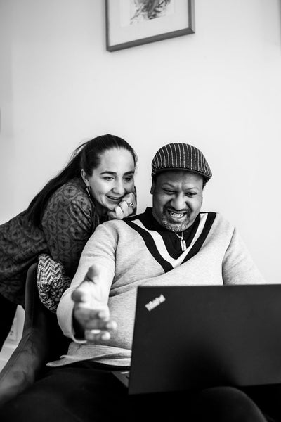 A smiling man sits with a laptop on his lap while a woman leans over his shoulder, also smiling, as they look at the screen together in a cozy indoor setting.