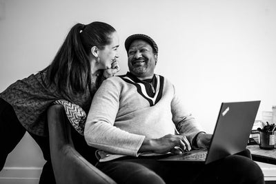 A woman leans in smiling at a man sitting with a laptop, both appearing happy and relaxed in a home setting. The image is in black and white.