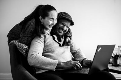 A woman hugs a smiling man from behind as he sits and types on a laptop. Both appear happy and are looking at the screen, sharing a joyful moment together in a cozy indoor setting.