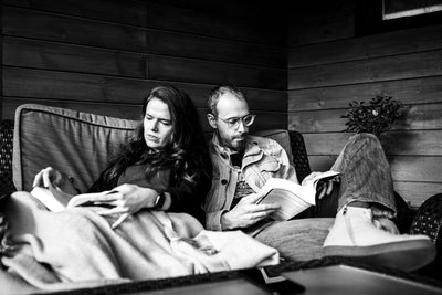 A woman and a man sit closely together on a sofa, both reading books. The scene is in black and white, with wooden paneling in the background and a blanket covering their laps.