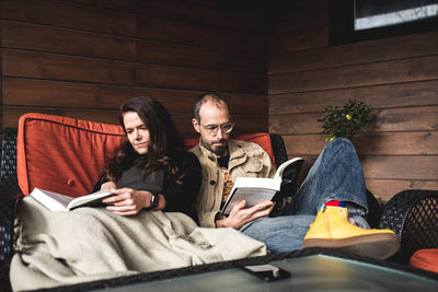 A woman and a man sit closely together on an outdoor couch, reading books. The woman is wrapped in a blanket and the man wears glasses and yellow boots. Wooden paneling and a small plant are in the background.