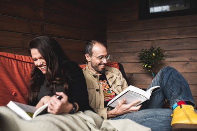 A woman and a man sit on an orange couch, laughing and reading books together. They are relaxed and cozy, with a blanket covering their legs and wooden walls in the background.