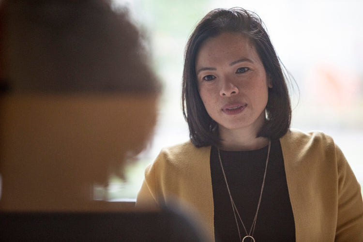 A woman with shoulder-length dark hair, wearing a yellow cardigan and a necklace, looks attentively at another out-of-focus person in the foreground during a conversation.