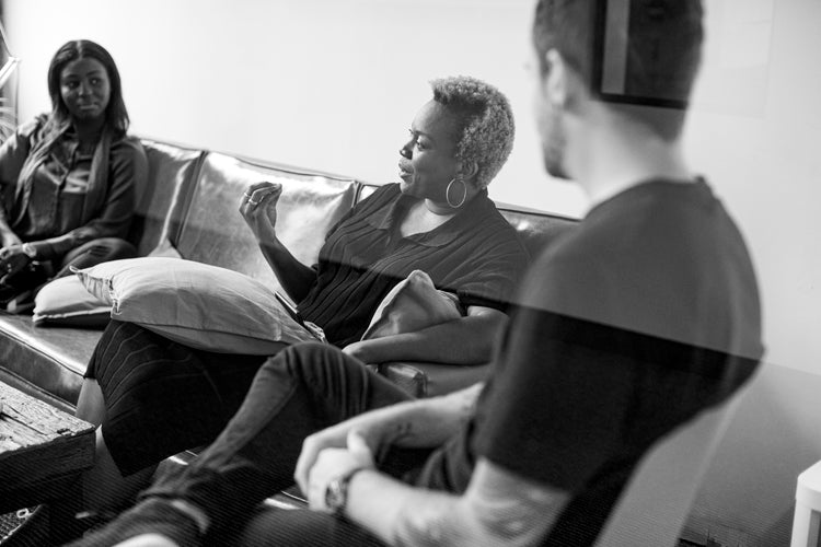 Three people sit on a couch having a conversation. The woman in the center gestures with her hand while talking, as the two others, a woman and a man, listen attentively. The scene appears casual and candid.