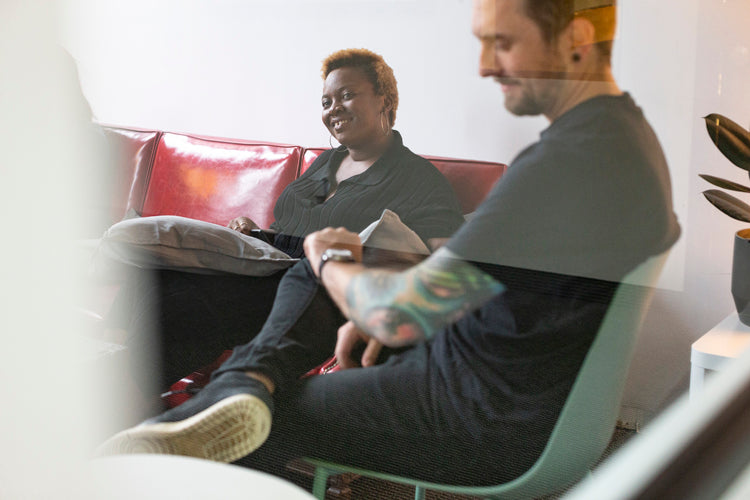 Two people sit on a red couch and a green chair, smiling and talking. One holds a pillow, and the other has tattoos on his arm. They appear relaxed in a casual, modern indoor setting.