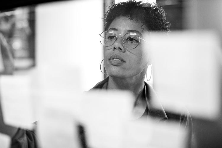 A woman with short curly hair and glasses looks thoughtfully at sticky notes on a glass wall, appearing focused and engaged in her work. The photo is in black and white.