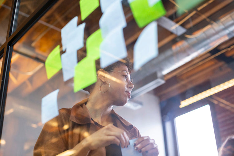 A woman stands behind a glass wall covered with sticky notes, looking to the side. She holds a sticky note in her hands. The background shows an office with exposed ceilings and natural light.
