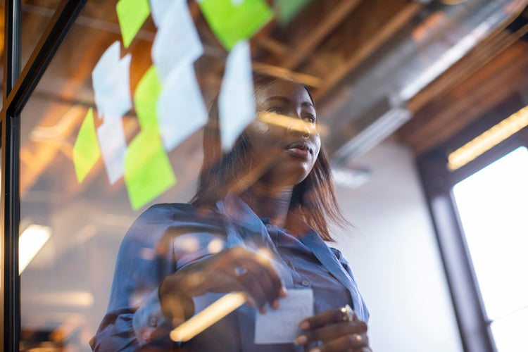 A woman stands behind a glass wall covered with colorful sticky notes, holding a note in her hand, appearing focused and thoughtful in a modern office setting with exposed beams and bright lighting.