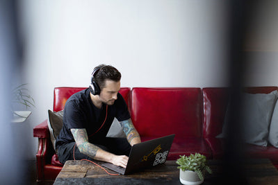 A person with tattoos, wearing headphones and a black shirt, sits on a red couch working on a laptop at a wooden coffee table. A small potted plant is on the table.