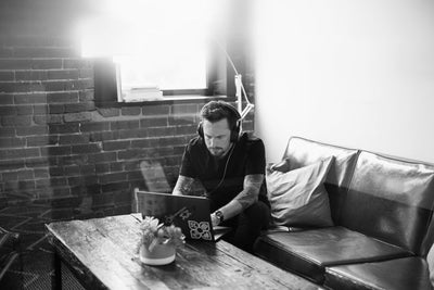A man wearing headphones sits on a couch, focused on a laptop placed on a wooden coffee table in front of him. The setting is a modern room with exposed brick walls and a small window.