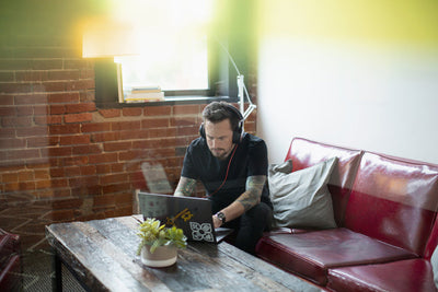 A man with tattoos sits on a red couch, wearing headphones and working on a laptop at a wooden coffee table with a small plant, in a room with brick walls and a window.