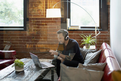 A man wearing headphones sits on a red sofa in a modern living room, smiling and gesturing at his laptop during a video call. There are plants on the wooden coffee table and a tall lamp in the background.