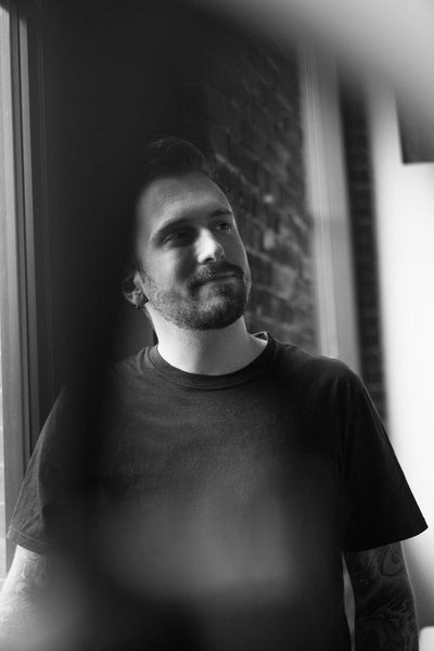 Black-and-white photo of a man with a beard and short hair, wearing a dark t-shirt, standing by a window and looking thoughtfully to the side. A blurred foreground element partially obscures the image.
