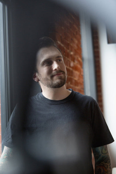 A man with a short beard and tattooed arms, wearing a black t-shirt, stands by a window with exposed brick behind him, looking thoughtfully to the side.