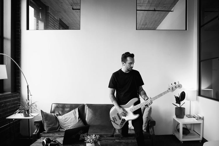 A man in a black t-shirt stands in a modern living room, playing an electric bass guitar. He is next to a couch with cushions, a laptop on the coffee table, and potted plants nearby. The room has large windows and high ceilings.