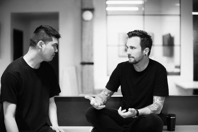 Two men in black shirts sit indoors, engaged in a serious conversation. One gestures with his hands while speaking, and the other listens attentively. The scene is in black and white.