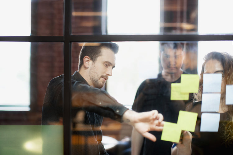 Three people stand behind a glass wall covered with sticky notes, discussing and pointing at the notes, collaborating in a bright, modern office space.