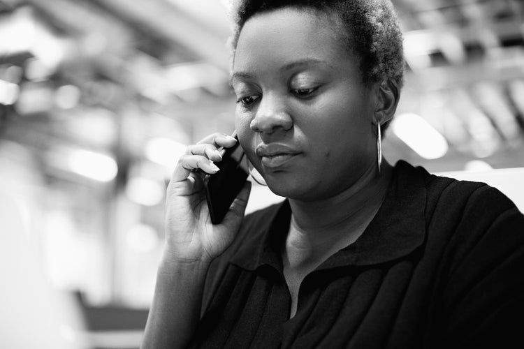 A woman with short curly hair is holding a phone to her ear, appearing focused while having a conversation. The background is blurred, suggesting an indoor office or work environment. The image is in black and white.