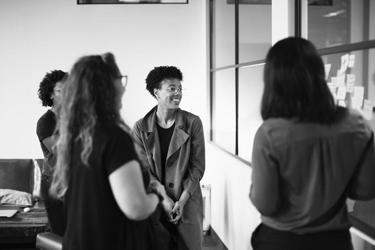 Four women stand and talk together in an office setting. One woman in a coat is smiling warmly at the group. Sticky notes are visible on the glass wall beside them. The image is in black and white.