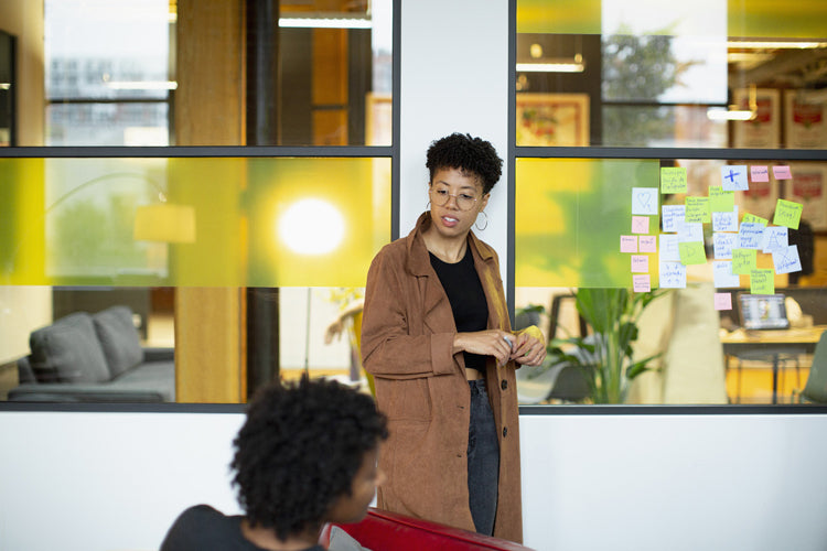 A person in a brown coat stands by a glass wall covered with colorful sticky notes, speaking to another person seated in the foreground. The setting appears to be a modern office with large windows.