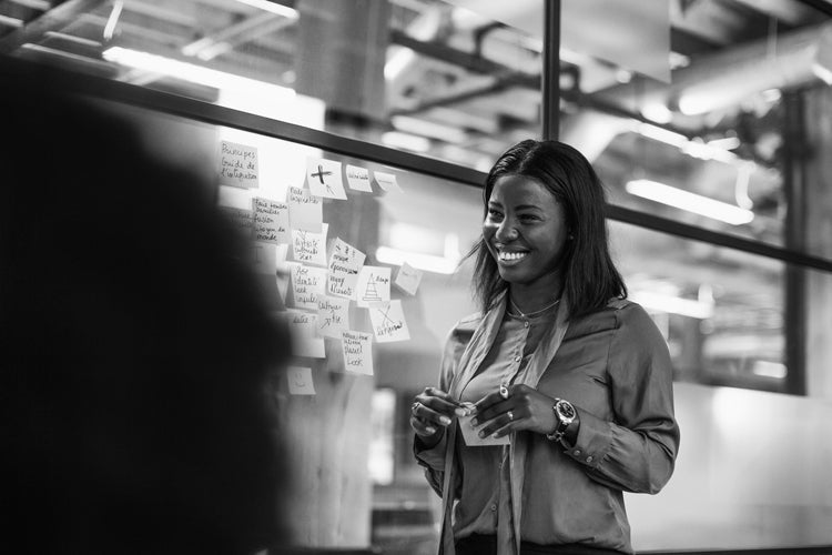 A woman smiles while standing near a glass wall covered with sticky notes in an office setting. She appears to be engaged in a discussion or presentation. The image is in black and white.