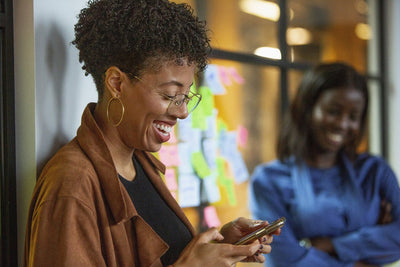 A woman wearing glasses and large hoop earrings smiles while using her phone, standing in front of a window with colorful sticky notes. Another woman in blue is smiling in the blurred background.
