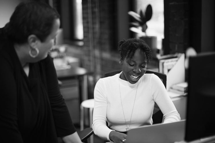 Two women are in an office. One woman is sitting at a desk, smiling while looking at a laptop. The other woman stands nearby, leaning in and engaging in conversation. The scene is in black and white.