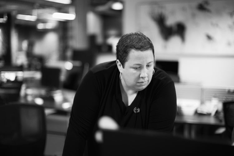 A person with short hair and a black top leans forward, looking intently at something off-camera, in a modern, blurred office setting.