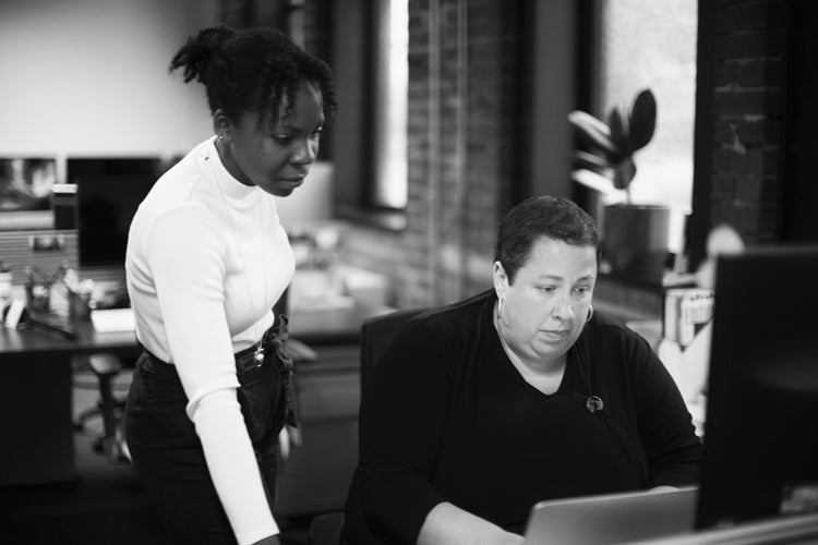 Two women in an office; one is seated and working on a computer while the other stands beside her, looking at the screen. The setting is modern with brick walls and large windows.