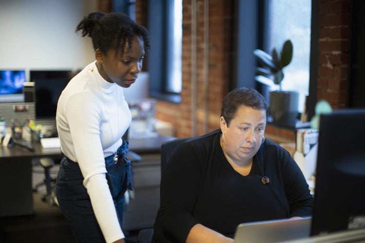 Two women in an office work together at a computer; one is standing and looking at the screen while the other is seated and focused on the monitor. The setting has brick walls and large windows.
