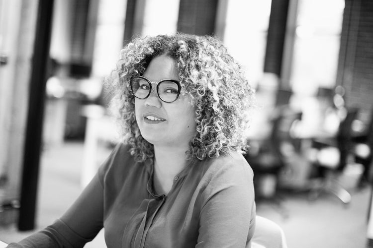 Black and white photo of a person with curly hair and glasses, smiling and sitting at a desk in a modern office environment with blurred background elements.
