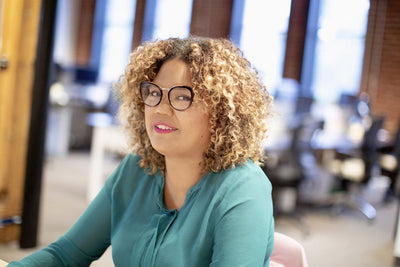 A woman with curly hair and glasses, wearing a green blouse, smiles while sitting in a modern office with blurred desks and windows in the background.