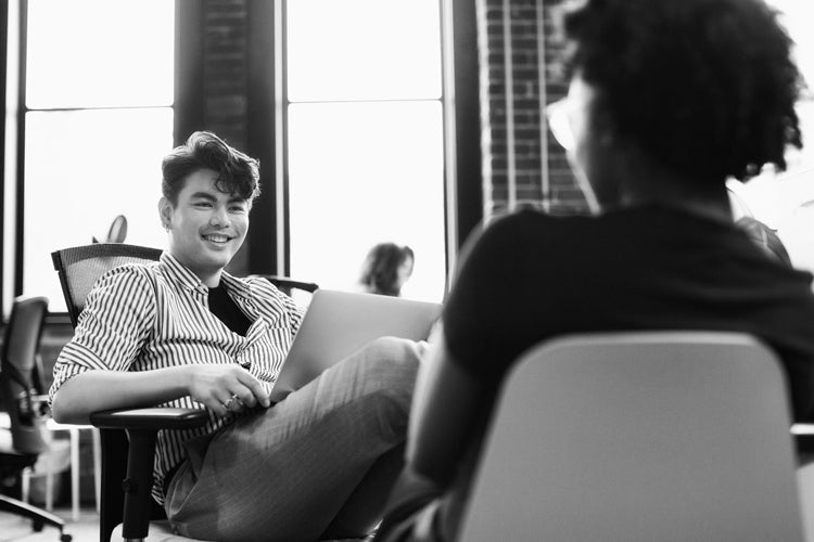 Two people sit across from each other in an office setting, one smiling while holding a laptop on their lap. Large windows and a brick wall are visible in the background. The image is in black and white.