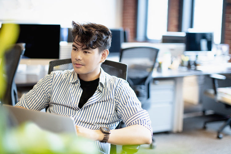 A person with short, styled hair sits at a desk in a modern office, looking to the side and smiling slightly. They are wearing a striped shirt over a black top. Computer monitors and desks are visible in the background.