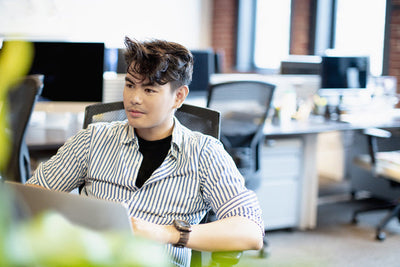 A person with short, styled hair sits at a desk in a modern office, looking to the side and smiling slightly. They are wearing a striped shirt over a black top. Computer monitors and desks are visible in the background.