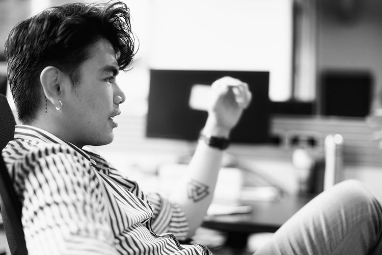 A person with short, styled hair and a striped shirt sits in an office chair, gesturing with one hand. The background shows a blurred desk, computer monitors, and office items. The image is in black and white.