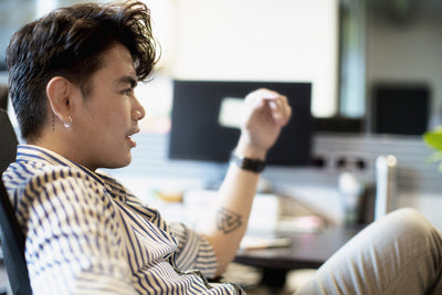 A young person with dark hair and a striped shirt sits in an office chair, talking animatedly. The background is blurred, showing a desk with a computer and various office items.