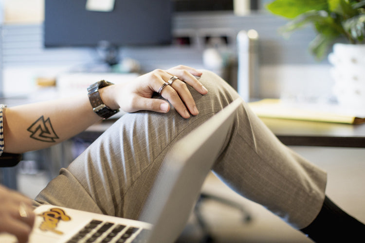 A person wearing light gray pants rests their hand on their crossed knee while working on a laptop. The individual has a geometric tattoo, wears rings, and sits in a modern office setting with plants in the background.