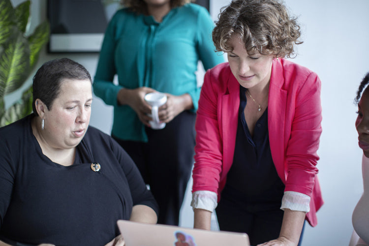 Two women are focused on a laptop, one seated and one standing in a pink blazer. Another person stands in the background holding a mug. They appear to be having a work discussion in an office setting.