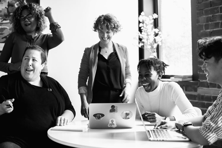 A group of five people sit and stand around a table with laptops, laughing and smiling together in a brightly lit office space.