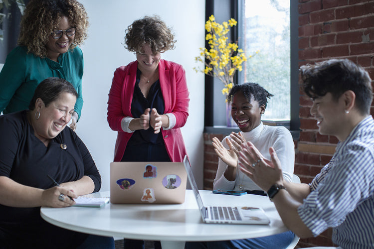 Five people gather around a table with laptops, laughing and clapping together in a bright room with exposed brick walls and a vase of yellow flowers, suggesting a joyful team meeting or collaboration.