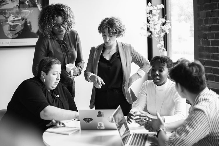 Five people are gathered around a table with laptops, engaged in a discussion. One person is standing and gesturing while others listen and interact, suggesting a collaborative work meeting in a modern office.