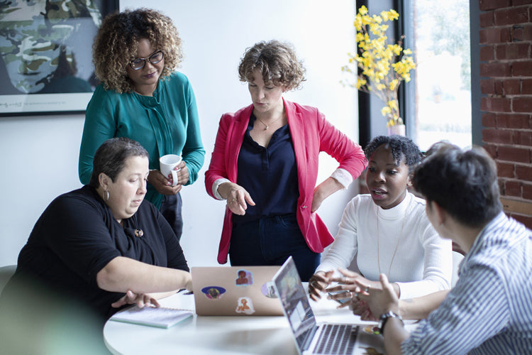Five people are gathered around a table with laptops, engaged in a discussion. Two are standing, and three are seated. The group appears focused and collaborative in a bright office with a large window and exposed brick wall.