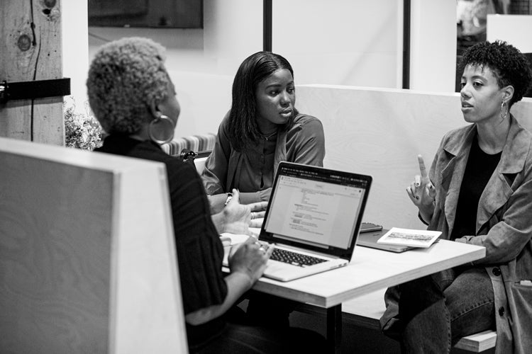 Three women sit at a table in an office, engaged in discussion. A laptop and notebook are on the table. One woman gestures with her hand as she speaks. The image is in black and white.