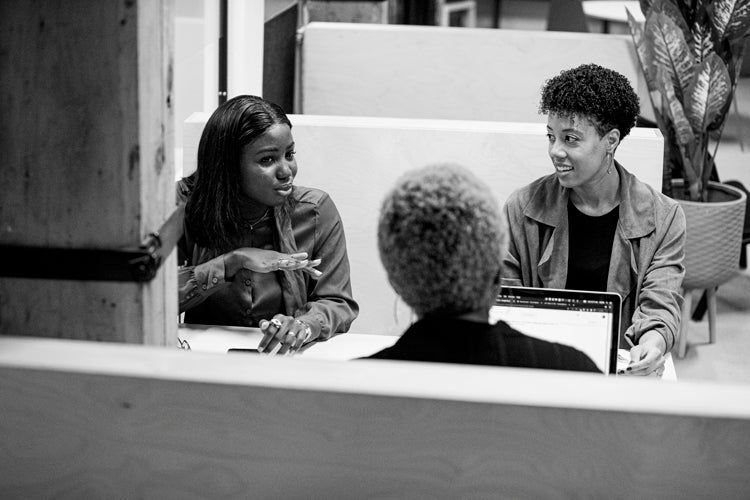 Three women sit at a table in an office setting, engaged in conversation. One woman gestures with her hands while speaking, and another smiles, listening attentively. A laptop is open in front of the third woman.