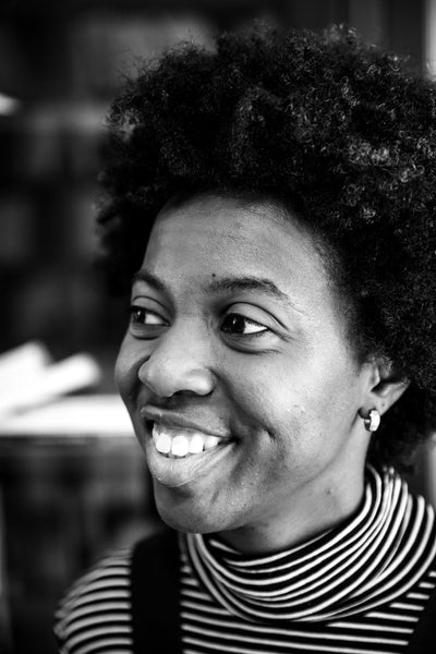 Black and white photo of a smiling person with curly hair, wearing a striped turtleneck and small hoop earrings, looking slightly to the side.