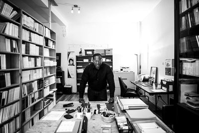A man stands leaning over a desk in a well-organized office filled with bookshelves, papers, and office supplies. The room is brightly lit, and the atmosphere appears focused and studious.
